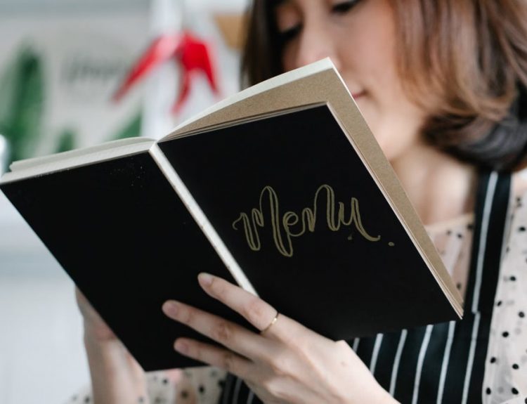 A chef in a polka dot blouse holds a menu book in a stylish restaurant, ready to present the dishes.
