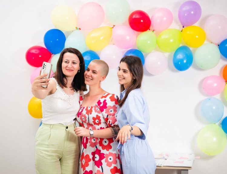 Three women smiling and taking a selfie against a vibrant balloon backdrop.