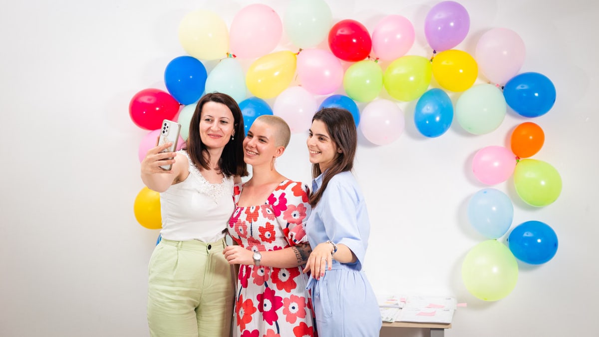 Three women smiling and taking a selfie against a vibrant balloon backdrop.